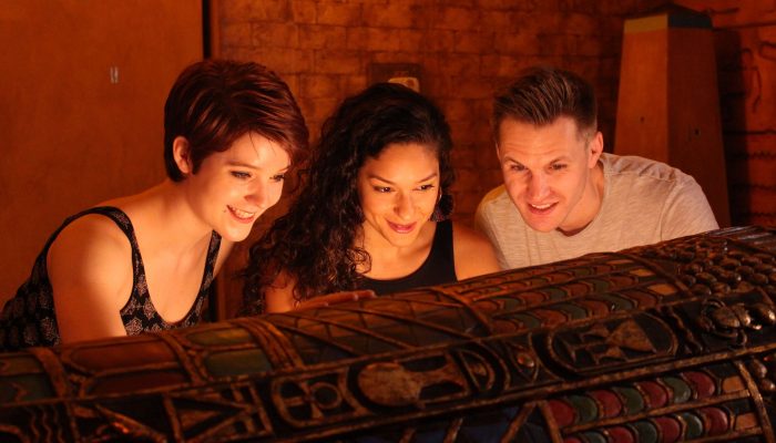 Three players in the egyptian tomb escape room looking down at a sarcophagus, their faces lit from below.